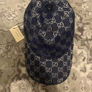 Gucci keps i blå denim, storlek M - Exklusiv keps från Gucci i blå denim med ikoniskt GG-mönster över hela kepsen. Klassisk böjd skärm och svart knapp på toppen. Insidan är fodrad i ljus bomull. Perfekt för dig som vill sticka ut med en lyxig accessoar.