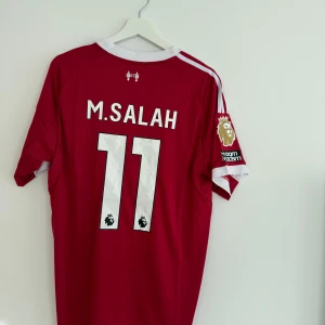 Liverpool M. Salah matchtröja röd - Säljer en officiell Liverpool FC fotbollströja med M. Salah och nummer 11 på ryggen. Tröjan är röd med vita detaljer, korta ärmar och Premier League-märke på ärmen. Materialet är lätt och andas, perfekt för träning eller match. Adidas-logga och Standard Chartered tryck på framsidan.