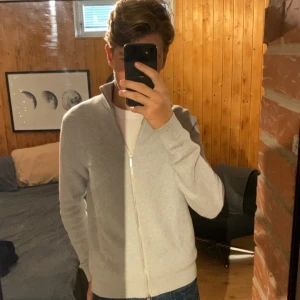 Stickad kofta från Selected Homme - Säljer en ljusgrå stickad kofta från Selected Homme. Koftan har hel dragkedja, ribbad krage och muddar samt en clean och stilren look. Perfekt för lager-på-lager och funkar till jeans eller chinos.