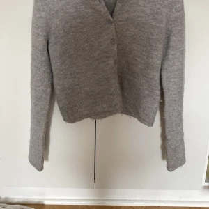 Grå stickad kofta från Gina Tricot - Mysig grå stickad kofta från Gina Tricot i storlek XS. Koftan har en enkel design med knappar framtill och långärmade ärmar. Materialet är mjukt och fluffigt, perfekt för kyliga dagar. Passformen är normal och färgen är ljusgrå, vilket gör den enkel att matcha med allt.