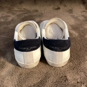 Vita sneakers från Philippe Model  - Snygga vita sneakers från Philippe Model med mörkblå häl och logotyp på sidan. Skorna har snörning och är tillverkade i skinn och mocka. Perfekta för dig som gillar stilrena och klassiska sneakers med en twist.  Skriv vid intresse och pris kan diskuteras.