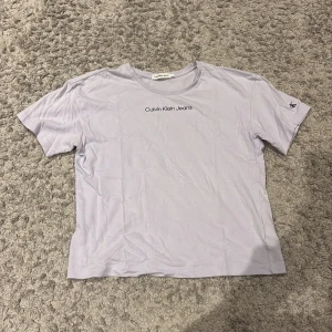 Ljuslila t-shirt från Calvin Klein Jeans - Snygg ljuslila t-shirt från Calvin Klein Jeans med svart logotyptryck på bröstet och ett litet 'ck' tryck på ärmen. Modellen är klassisk med rund hals och korta ärmar