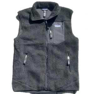 Svart fleeceväst från Patagonia - Mysig svart fleeceväst från Patagonia med hög krage och hel dragkedja framtill. Västen har två sidofickor med dragkedja och en bröstficka med logga. Perfekt för lager-på-lager och outdoor-vibe.