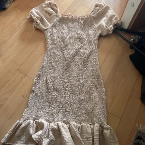 Beige kort klänning med volang - Supersöt beige kortklänning med puffärmar och volang nertill. Smockad överdel ger en tight passform och de korta ärmarna har en romantisk känsla. Perfekt för sommarens alla tillfällen!