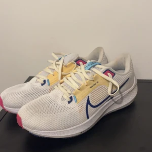 Nike Pegasus 40 sneakers vit/blå/gul - Nike Pegasus 40 sneakers i vitt mesh med blå swoosh, gula och blå detaljer samt rosa inslag. Skorna har snörning, platt sula och ventilerande ovandel. Perfekta för löpning eller träning, med en modern och sportig look.