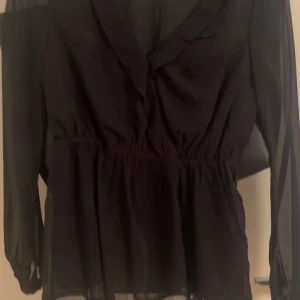 Svart omlottblus med knytband - Snygg svart blus från Nelly med v-ringning och långärmade transparenta ärmar. Perfekt för dig som gillar stilrena plagg med en twist.  använd en gång på nyår.