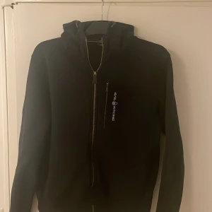 Svart Sail Racing hoodie med dragkedja - Svart hoodie från Sail Racing i storlek M. Tröjan har hel dragkedja framtill, en bröstficka med dragkedja och Sail Racing-logga på bröstet. Mjuk insida och huva för extra komfort. 
