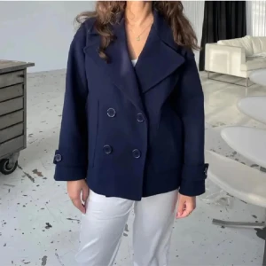 Mörkblå kappa Design by Si - Säljer en snygg mörkblå kappa med dubbelknäppning och breda slag. Jackan hae inga större tecken på användning och har använts sparsamt.