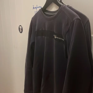 Marinblå Sail Racing sweatshirt - Marinblå långärmad sweatshirt från Sail Racing med diskret logga och svart band över bröstet. Klassisk rund halsringning med vit detalj. Perfekt för dig som gillar stilren och sportig look.