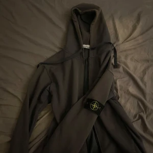 Mörkgrå hoodie från Stone Island - Säljer en mörkgrå hoodie från Stone Island med dragkedja och klassisk logga på ärmen. Tröjan har huva med snörning och långa ärmar. Perfekt för dig som gillar streetwear och vill ha något stilrent men ändå med edge.