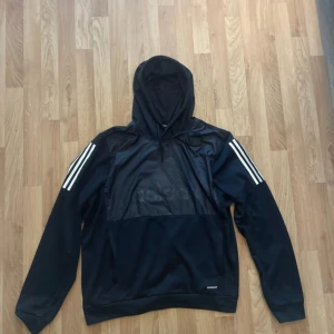 Svart hoodie från Adidas XL - Svart hoodie från Adidas i storlek XL med klassiska vita ränder på axlarna. Tröjan har huva med snörning och är tillverkad i polyester med meshdetaljer på insidan. Perfekt för sport eller chill, med långärmad passform och stilren look.