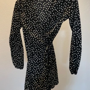 Svart prickig omlottklänning Boohoo - Snygg svart kort omlottklänning från Boohoo med vita prickar. Klänningen har långa ärmar, v-ringad hals och knyt i midjan för en smickrande siluett. Perfekt för dig som gillar trendiga mönster och en feminin look.