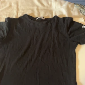 Svart t-shirt från Calvin Klein Jeans - Svart t-shirt från Calvin Klein Jeans med klassisk rund hals och korta ärmar. Diskret CK-logga i vitt på ena ärmen ger en clean och stilren vibe. Mjuk bomullskvalitet som är skön mot huden och passar perfekt till jeans eller shorts.