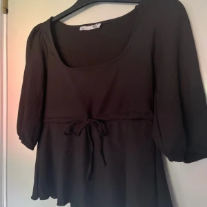 Svart blus med knyt från lager 157 - Supersöt svart blus från lager 157 med trekvartsärmar och knyt i midjan. Blusen har en fyrkantig ringning och är tillverkad i ett ribbat material som ger en snygg struktur. Perfekt för dig som gillar en avslappnad stil.