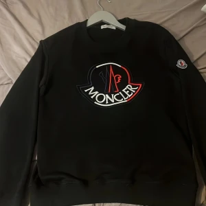 Svart Moncler sweatshirt med stor logga - Svart sweatshirt från Moncler med stor broderad logga i vitt, rött och blått på bröstet. Klassisk rund halsringning och Moncler-märke på ärmen. Perfekt för dig som vill ha en clean men ändå lyxig look.