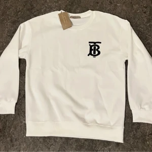 Vit sweatshirt från Burberry - Vit sweatshirt från Burberry med svart TB-logga på bröstet. Klassisk rund halsringning och ribbade muddar vid ärmslut och nederkant. Tröjan är i bomull och har en clean, stilren look som passar perfekt till jeans eller dressade byxor.