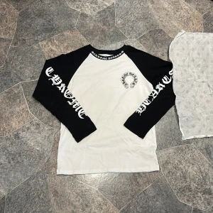 Chrome Hearts svartvit långärmad tröja - Säljer en långärmad tröja från Chrome Hearts med svartvita kontraster. Tröjan har svarta ärmar med tryck och stor logga både fram och bak. Rund hals med tryckt text runt kragen. Perfekt för dig som gillar streetwear och vill sticka ut.