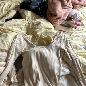 Ribbad beige långärmad topp NA-KD - Säljer en ribbad beige långärmad topp från NA-KD i storlek XS. Toppen har en snygg, djup rundad ringning och är figurnära i modellen. Perfekt att styla med jeans eller kjol för en clean look.