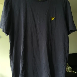Mörkblå t-shirt från Lyle & Scott - Snygg mörkblå t-shirt från Lyle & Scott med klassisk gul logga på bröstet. Enkel och stilren design med rund hals och korta ärmar. Perfekt för dig som gillar en clean look och vill ha ett plagg som funkar till det mesta.