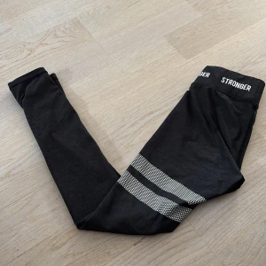 Svarta leggings från Stronger - Mörka gråa leggings från Stronger med vita prickiga ränder runt ena benet och logga i midjan. De är tighta och har hög midja, perfekta för träning eller chill. Materialet är stretchigt och mjukt, vilket gör dem riktigt sköna att ha på sig.