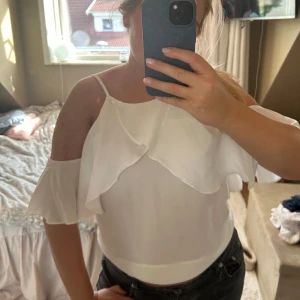 Vit blus med volang från Crocker - Supersöt vit blus från Crocker med tunna axelband och offshoulder-ärmar. Blusen har volangdetaljer framtill och är tillverkad i ett lätt och luftigt material som passar perfekt till varma dagar.