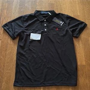 Svart Polo Ralph Lauren t-shirt S - Svart t-shirt från Polo Ralph Lauren i klassisk pikéstil med krage och tre vita knappar framtill. Liten röd broderad logga på bröstet. T-shirten är kortärmad och gjord i mjuk bomull som känns skön mot huden. Perfekt för en clean och stilren look.