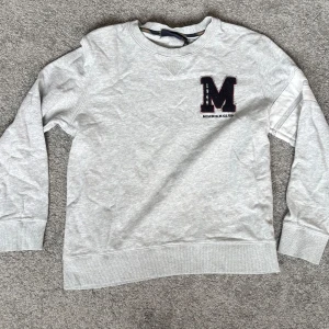 Grå sweatshirt från Morris R. Club - Snygg ljusgrå sweatshirt från Morris R. Club med broderad svart M och årtal på bröstet. Klassisk rund halsringning, ribbade muddar och lång ärm. Perfekt för en chill och sportig look.