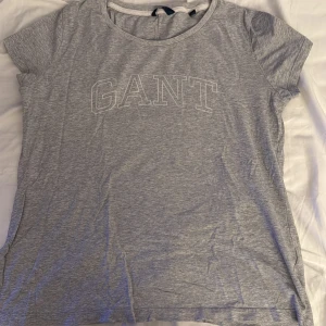 Grå t-shirt från GANT med tryck - Snygg grå t-shirt från GANT i mjuk bomull. Klassisk rund hals och korta ärmar. Framsidan har ett stilrent GANT-tryck i vitt. Perfekt för chill dagar och enkel att matcha med jeans eller shorts.