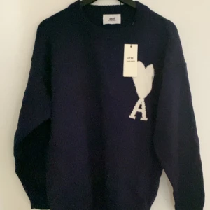 Mörkblå stickad tröja från Ami (storlek S) - Snygg mörkblå stickad tröja från Ami Alexandre Mattiussi med vit hjärtlogga och bokstaven A på bröstet. Tröjan har rund halsringning, ribbade muddar och lång ärm. Perfekt för dig som gillar stilrena och trendiga plagg.