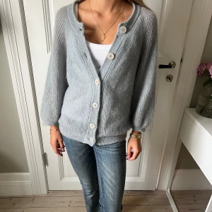 Ljusgrå/blå kofta från Filippa K - Supermjuk och fluffig ljusgrå kofta från Filippa K i stickad ullmix. Koftan har stora vita knappar framtill, v-ringad hals och lite ballongformade ärmar. Perfekt att slänga över ett linne eller t-shirt för en chill look.