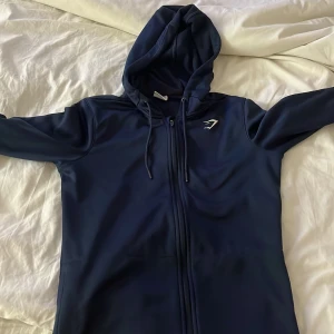 Mörkblå hoodie från Gymshark - Säljer en mörkblå hoodie från Gymshark med dragkedja och snörning i huvan. Tröjan har lång ärm och ett stilrent Gymshark-logo på bröstet. Perfekt för chill eller träning, mjukt material och snygg passform.