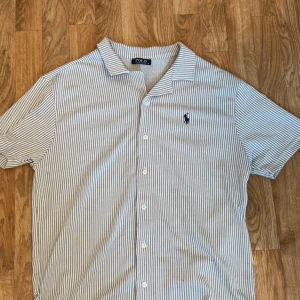 Ralph Lauren skjorta - Snygg kortärmad skjorta från Polo Ralph Lauren i vitt och grått med tunna vertikala ränder. Klassisk krage, knäppning framtill och broderad logga på bröstet. Perfekt för en clean och stilren look. Endast använd ett par gånger.
