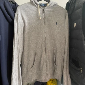 Grå hoodie från Polo Ralph Lauren - Grå hoodie från Polo Ralph Lauren med dragkedja framtill, huva med snörning och två fickor. Klassisk blå logga broderad på bröstet. Tillverkad i mjuk bomullsmix, perfekt för chill dagar. Långärmad och normal passform.