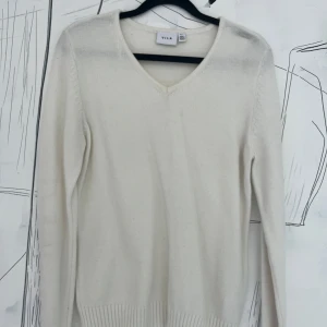 Vit v-ringad stickad tröja från Vila - Klassisk vit stickad tröja från Vila med v-ringning och långa ärmar. Tröjan har ribbade muddar vid ärmslut och nederkant, vilket ger en snygg och enkel look. Perfekt att matcha med jeans eller kjol för en clean stil.