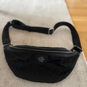 Svart magväska från DKNY - Snygg svart magväska från DKNY med justerbart band och silverfärgad dragkedja. Väskan har diskret DKNY-mönster över hela ytan och ett metallogo framtill. Invändigt finns ett fack med dragkedja för extra förvaring. Perfekt för att bära det viktigaste på stan.