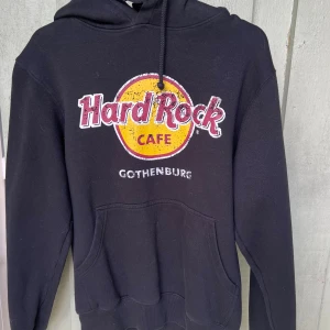 Svart Hard Rock Cafe hoodie M - Svart hoodie från Hard Rock Cafe Gothenburg med klassisk logga i gult och rött framtill. Tröjan har huva med snörning och stor magficka. Tillverkad i mjuk bomull och polyester, perfekt för en chill och avslappnad stil.