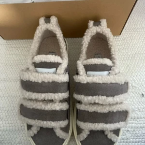 Grå sneakers från Inuikii med fårull - Unika grå sneakers från Inuikii med fluffiga detaljer i beige fårull och breda kardborreband. Skorna har en chunky vit sula och är tillverkade i mocka och fårull, perfekta för dig som vill ha något extra mysigt och trendigt.💞