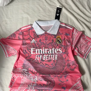 Real Madrid rosa fotbollströja Adidas M - Snygg Real Madrid fotbollströja från Adidas i rosa med grått drakmönster över hela tröjan. Klassisk vit krage och vita detaljer på ärmarna. Broderat klubbmärke på bröstet och tryckt sponsorlogga. Tillverkad i lätt och ventilerande polyester.
