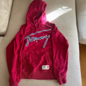 Rosa hoodie från Tommy Jeans XS - Säljer en snygg rosa hoodie från Tommy Jeans i storlek XS. Tröjan har huva med snörning, stor broderad logga framtill i blått och vitt samt klassisk känguruficka. Perfekt för dig som gillar streetstyle och vill sticka ut.