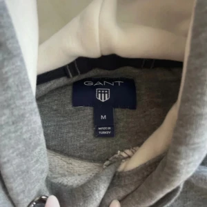 Grå hoodie från GANT med tryck - Snygg grå hoodie från GANT med stort vitt tryck på bröstet och coola förstärkningar på armbågarna. Tröjan har huva med snörning och ribbade muddar. Perfekt för dig som gillar sportig och avslappnad stil.