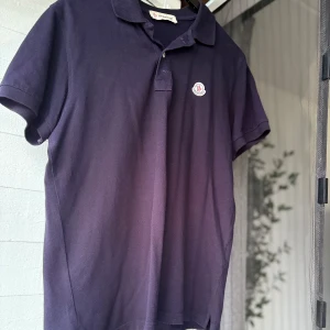 Mörkblå Moncler pikétröja - Snygg mörkblå piké från Moncler med klassisk krage och två knappar framtill. T-shirten har korta ärmar och ett broderat Moncler-emblem på bröstet. Tillverkad i mjuk bomull som känns skön mot huden. Perfekt för dig som gillar stilrena och exklusiva plagg. (Den är i bra skick men saknar en kanp alltså den som stänger vid halsen annars är den bra) 