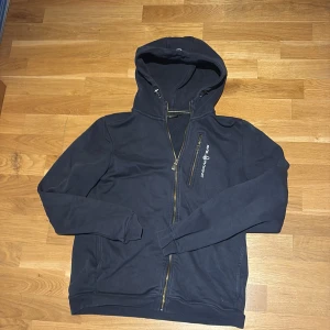 Mörkblå hoodie från Sail Racing - Mörkblå hoodie från Sail Racing med dragkedja framtill och bröstficka med dragkedja. Tröjan har huva med snörning och är tillverkad i mjuk bomull. Perfekt för dig som gillar sportig och stilren look.