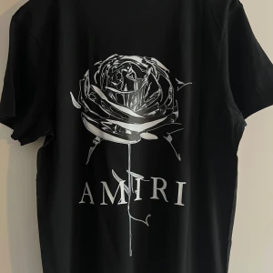 Svart Amiri t-shirt med tryck - Svart t-shirt från Amiri med coolt tryck på både fram- och baksidan. Framsidan har Amiri-loggan med färgstänk och baksidan har en stor silverros med logotyp. Klassisk rund hals och korta ärmar. Materialet är mjuk bomull som känns skönt mot huden.