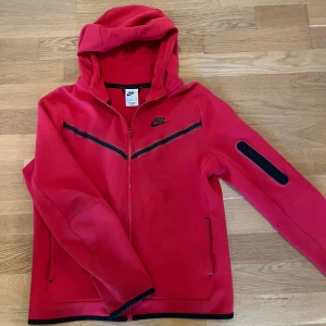 Nike tech fleece  - Nike vindjacka i rött med svarta detaljer och dragkedja. Jackan har huva, svarta ränder över bröstet och en ficka på ärmen. Perfekt för dig som gillar sportig stil och vill ha något bekvämt och snyggt till vardags.
