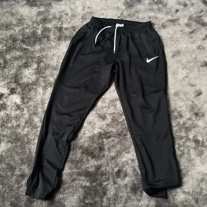 Svarta Nike Dri-FIT träningsbyxor M - Svarta träningsbyxor från Nike med Dri-FIT-teknologi och vit snörning i midjan. Byxorna har en smal passform och elastisk midja, samt Nike-logga på låret. Perfekta för sport eller chill. Materialet är lätt och andas.
