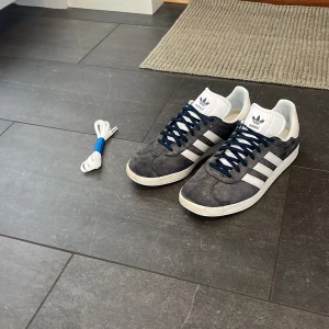 Adidas Gazelle mörkblå sneakers mocka - Säljer ett par Adidas Gazelle sneakers i mörkblå mocka med vita detaljer och klassiska tre ränder på sidorna. Skorna har vit plös och häl, mörkblå snörning samt medföljande extra vita skosnören. Skorna är aningen solblekta men bortsett från det har skorna inga defekter. Skriv gärna vid frågor eller funderingar. Pris kan diskuteras!