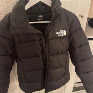 Svart pufferjacka från The North Face - Säljer en svart pufferjacka från The North Face med klassisk logga på bröstet och ryggen. Jackan har hög krage, dragkedja framtill och är vadderad för extra värme. Nypris 2500kr