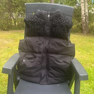 Svart dunväst Glorious Gangsta - Snygg svart dunväst från Glorious Gangsta med fet logga på bröstet och coolt geometriskt mönster upptill. Västen har hög krage, dragkedja framtill och två stora fickor med zip. Materialet är polyester och dun, perfekt för kyliga dagar. Om du vill se mer bilder skriv till mig