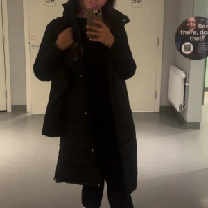 Svart lång dunjacka med huva - Säljer en svart lång dunjacka med huva och hög krage. Jackan har quiltad design och stängs med både dragkedja och knappar. 