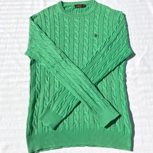 Grön kabelstickad tröja från Morris - Snygg grön kabelstickad tröja från Morris i storlek M och 100% pima bomull. Tröjan har rund halsringning, ribbade muddar och ett broderat emblem på bröstet. Perfekt för dig som gillar klassisk stil med en twist av färg. Tröjan är i mycket bra skick och har endast används 3-5 gånger utan några fläckar eller imperfektioner. 